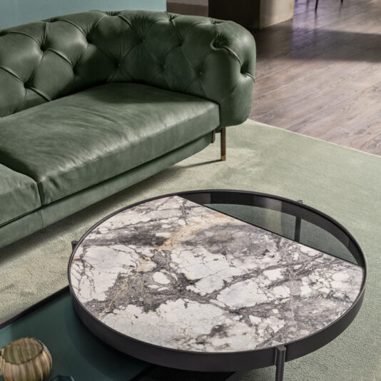 The cozy living room showcases a Modern Low Button Upholstered Suede Sofa with a stylish rounded backrest, paired with a round coffee table featuring a split design—half marble and half dark metal. The setup is completed by placing the table on a pale green rug that rests over a wooden floor.