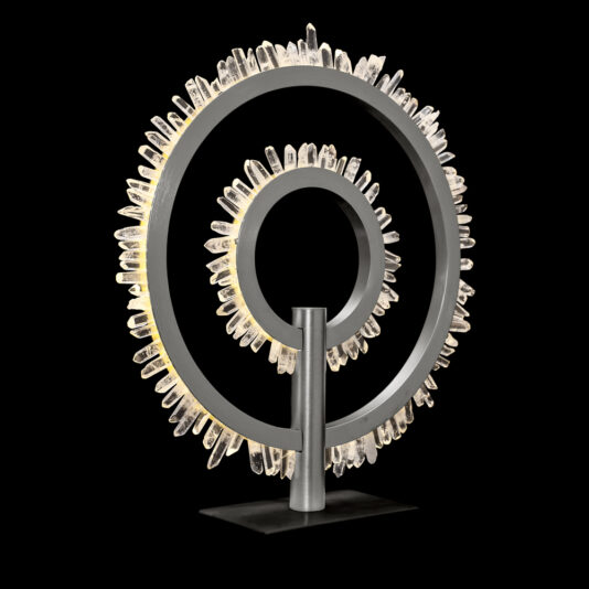 The Modern Quartz Crystal Hoop Nickel Table Lamp resembles a modern art sculpture with two concentric rings: a smaller inner ring and a larger outer ring. Both rings are adorned with clear quartz crystal formations along their circumferences, and the piece is set against a black background that highlights its intricate design.