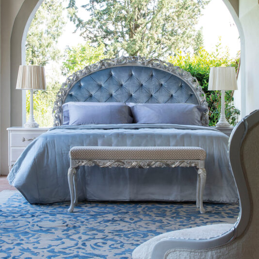 Luxurious bedroom featuring an Ornate Carved Button Upholstered Bed in silver, adorned with matching light blue bedding and complemented by elegant bedside tables with lamps. A decorative bench sits at the foot of the bed, while a grand archway offers a lush outdoor view.