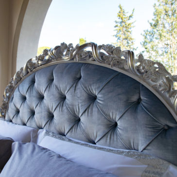 Ornate Carved Button Upholstered Bed A close-up of the Ornate Carved Button Upholstered Bed, featuring intricate silver floral carvings and blue button-tufted upholstery. The bed is adorned with plush pillows and sits near a large window that provides a view of trees and a bright sky.