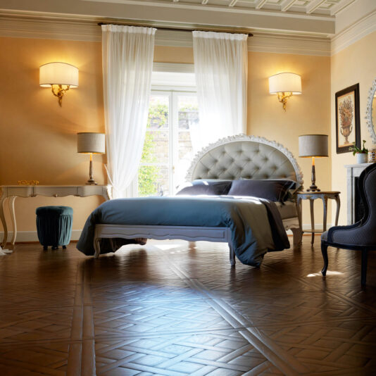 A luxurious bedroom features an Ornate Carved Rose Button Upholstered Bed adorned with plush pillows and a blue-gray comforter. Two bedside tables with matching lamps flank the bed, complemented by wall-mounted sconces. Large windows covered in sheer white curtains let in natural light, while intricate wood patterns decorate the floor.