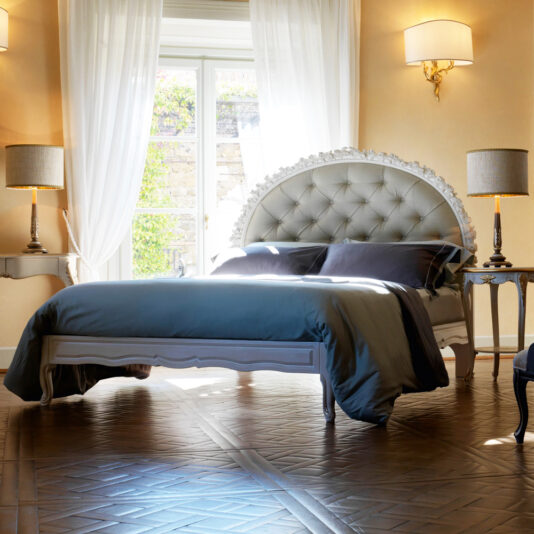 A luxurious bedroom features an Ornate Carved Rose Button Upholstered Bed with a tufted headboard, dressed in dark blue bedding. Matching nightstands with elegant lamps flank the bed. Light pours in from the large window behind, adorned with sheer curtains. The rich wood flooring completes the sophisticated look.