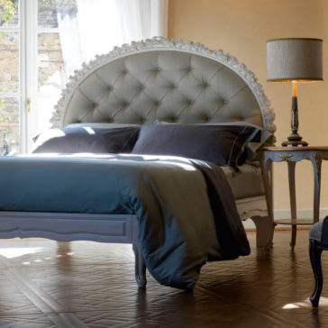 Ornate Carved Rose Button Upholstered Bed A well-lit bedroom showcases the Ornate Carved Rose Button Upholstered Bed, adorned with a plush, tufted headboard and dressed in dark bedding. Sunlight streams in through a window on the left. A vintage-style nightstand with an elegant lamp sits next to the bed, adding a cozy ambiance to the space.