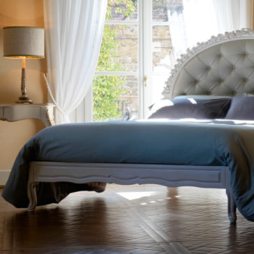 Ornate Carved Rose Button Upholstered Bed A brightly lit bedroom featuring the Ornate Carved Rose Button Upholstered Bed with blue bedding. A lamp sits on an elegant side table beside the bed. White curtains hang from large windows, allowing natural light to flood the room. The floor is wooden.