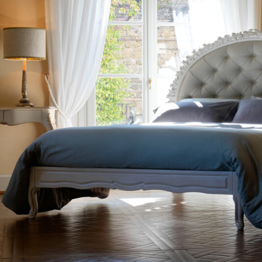 A brightly lit bedroom featuring the Ornate Carved Rose Button Upholstered Bed with blue bedding. A lamp sits on an elegant side table beside the bed. White curtains hang from large windows, allowing natural light to flood the room. The floor is wooden.