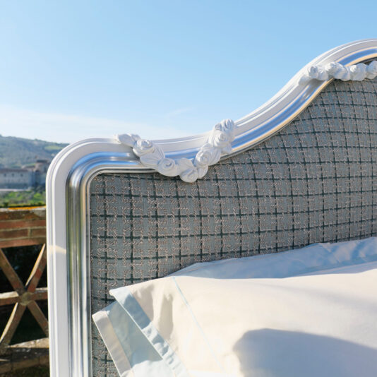 Close-up of the Ornate Classic Style Bed's headboard, showcasing a white and silver frame adorned with intricate floral details. The headboard is upholstered in textured gray fabric, complemented by a light blue pillow with white trim. The backdrop reveals an outdoor scene with distant buildings.