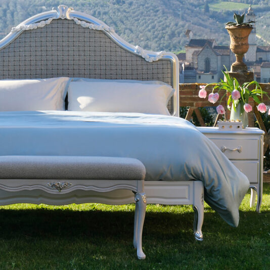 The outdoor scene showcases the **Ornate Classic Style Bed** adorned with light blue bedding and white pillows, complete with an intricately designed headboard. A matching bench and nightstand are situated close by. In the background, rolling green hills and a segment of a historic building set the picturesque scenery, while pink flowers enhance the overall ambiance.