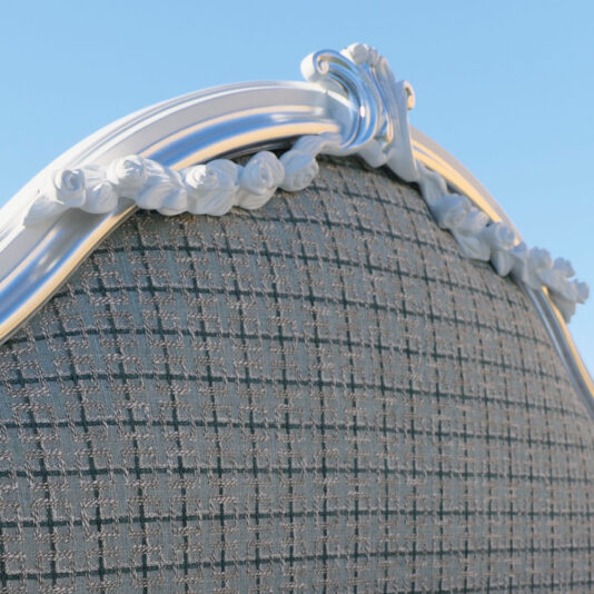 A close-up of the Ornate Classic Style Bed's headboard showcases a textured grey fabric pattern, highlighted by an elegant silver trim and intricate decorative rose carvings at the top, all set against a backdrop of a clear blue sky.