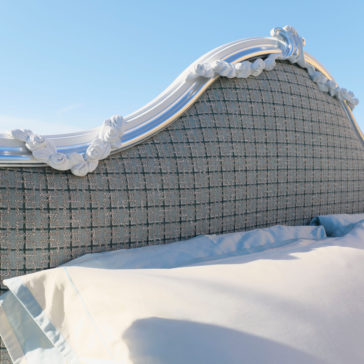 The image showcases a close-up of the Ornate Classic Style Bed headboard, featuring gray, textured fabric with exquisite white floral detailing along the top edge. A portion of light blue and white bedding is visible in the foreground, with a clear blue sky providing a scenic backdrop.