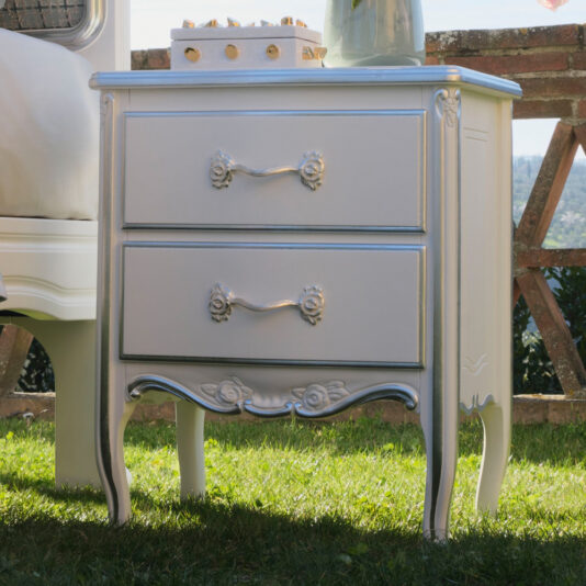 The Ornate Classic Style Bedside Cabinet is a white, vintage-style bedside table that features two drawers with elaborate handles and intricately carved floral details. It stands on elegantly curved legs and is pictured outdoors on grass, against a brick wall with lush greenery in the background.