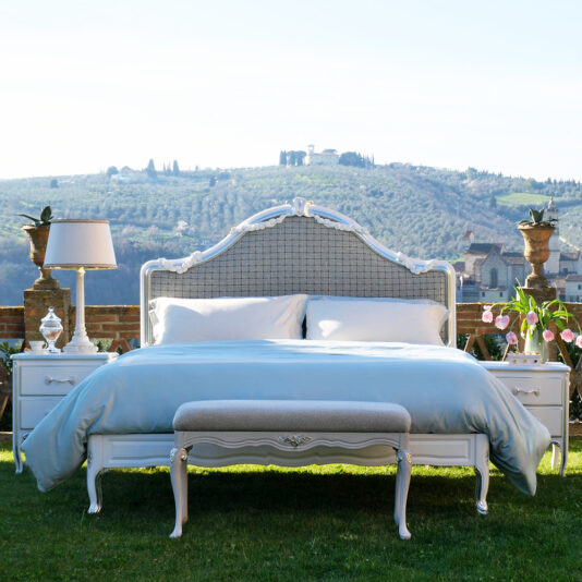 An elegant white-framed bed adorned with luxurious light blue bedding is positioned outdoors on a grassy lawn. Accompanying the bed are Ornate Classic Style Bedside Cabinets featuring decorative lamps and potted plants. The backdrop includes rolling green hills dotted with houses and trees.