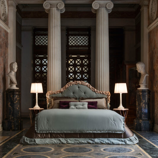 A luxurious bedroom showcasing the Ornate Classic Style Button Upholstered Bed, covered in elegant light blue bedding and flanked by two bedside tables with white lamps. The room features classic elements like columns and marble busts on pedestals, creating a grand aesthetic.