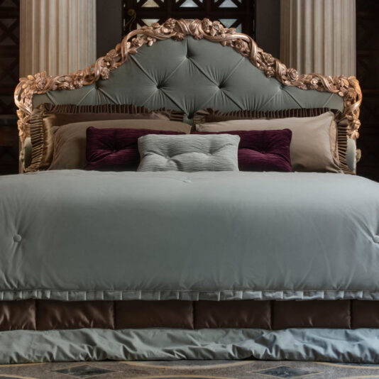 The Ornate Classic Style Button Upholstered Bed features a luxurious, button-upholstered design with an intricately carved headboard in gold. The bed is adorned with multiple pillows in shades of purple, gray, and beige, and is covered with a light gray comforter. The opulent setting includes classic columns in the background.