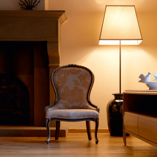 Ornate Venetian Style Occasional Chair An Ornate Venetian Style Occasional Chair with a vintage floral pattern is placed in a warmly lit room beside a fireplace. A tall floor lamp casts a soft glow nearby, illuminating a dark decorative vase and a table adorned with a white ceramic bird ornament.
