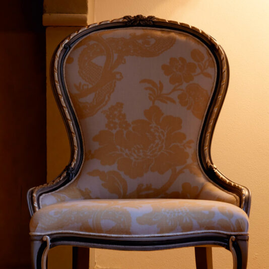 Ornate Venetian Style Occasional Chair An Ornate Venetian Style Occasional Chair with an intricate floral upholstery pattern in soft beige and gold tones is centered against a warmly lit background. The chair features a curved wooden frame and a plush seat, imparting an elegant and classic look.