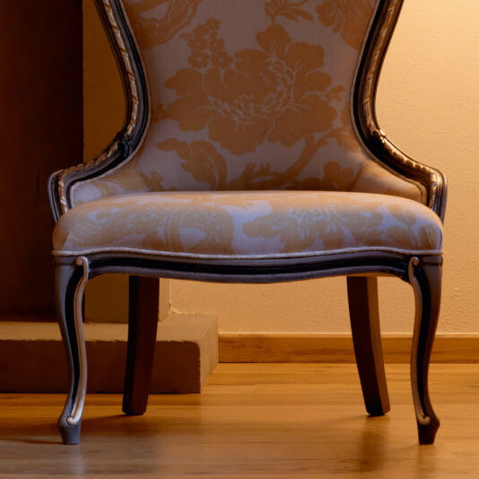 Ornate Venetian Style Occasional Chair A close-up view of the Ornate Venetian Style Occasional Chair showcases its upholstered design adorned with a floral pattern. The chair features wooden legs and elegant carvings along the edges, positioned on a wooden floor with a softly-lit background, creating a warm and inviting ambiance.