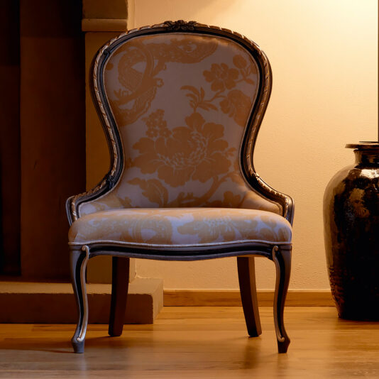 An Ornate Venetian Style Occasional Chair with a floral upholstery design sits in a softly lit room. The chair features a high, rounded back and curved wooden legs. To the right of the chair, a large decorative ceramic vase stands on the wooden floor. The background showcases a beige wall.