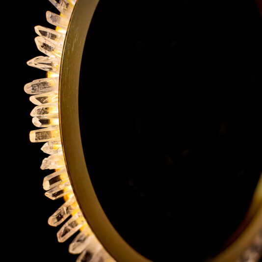 Large Quartz Crystal Gold Round Chandelier A close-up photo of a circular metallic object, likely a mirror frame, edged with illuminated crystal-like structures against a black background. The crystals are evenly spaced around the frame and glow warmly, creating a striking contrast with the dark surroundings.