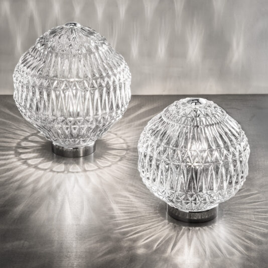 Two Round Murano Glass Table Lamps with intricate geometric patterns rest on a metallic surface. The lamps emit a soft light, casting decorative shadows and reflections on the surface and the wall behind them. The bases of the lamps are made of metal.