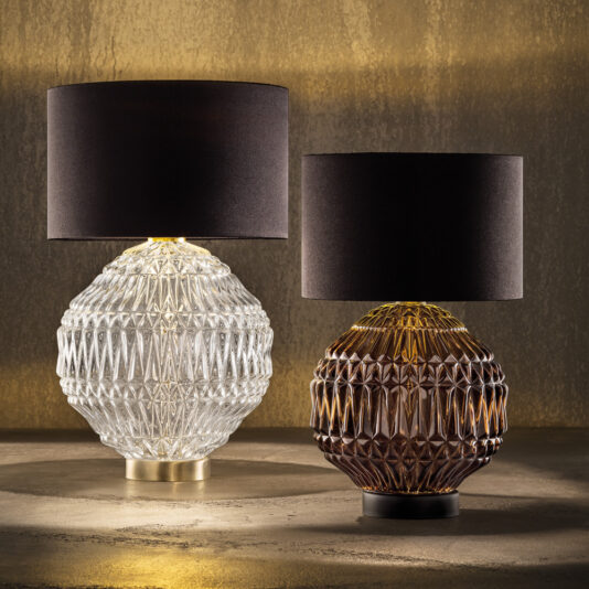 Two intricately designed Round Murano Glass Table Lamps With Shades create a captivating display. The left lamp features a clear glass base with a dark cylindrical shade, while the right boasts an amber-colored base with the same style of shade. These lamps are set against a warm, textured background.