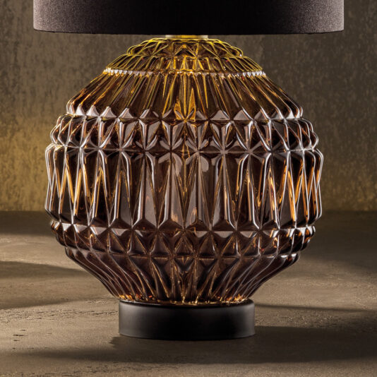 Round Murano Glass Table Lamp With Shade A close-up of the Round Murano Glass Table Lamp With Shade reveals its dark, circular silhouette adorned with intricate geometric patterns. The amber base is decorated with a starburst design that catches and reflects light, creating a warm and elegant ambiance.