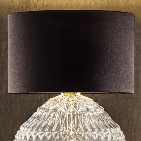Round Murano Glass Table Lamp With Shade A close-up of the Round Murano Glass Table Lamp With Shade reveals its textured, clear base and cylindrical, black lampshade. The lamp is turned on, emitting a soft glow that accentuates the intricate design of the glass base against a warm, neutral background.