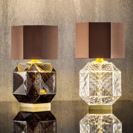 Two geometric table lamps with brown, angular lampshades. The left features a dark, faceted base and the right a clear, faceted base reminiscent of the Small Murano Glass Table Lamp. Both lamps stand on reflective surfaces against a textured beige wall.