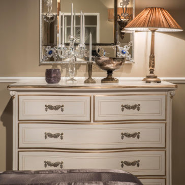Tall Classic Louis Style Chest Of Drawers The Tall Classic Louis Style Chest of Drawers, elegantly cream-colored with golden accents, is adorned with decorative candle holders, a reflective silver bowl, and a lit table lamp with a pleated shade. A large, ornate mirror hangs above the dresser, reflecting part of the room.