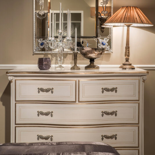 The Tall Classic Louis Style Chest of Drawers, elegantly cream-colored with golden accents, is adorned with decorative candle holders, a reflective silver bowl, and a lit table lamp with a pleated shade. A large, ornate mirror hangs above the dresser, reflecting part of the room.