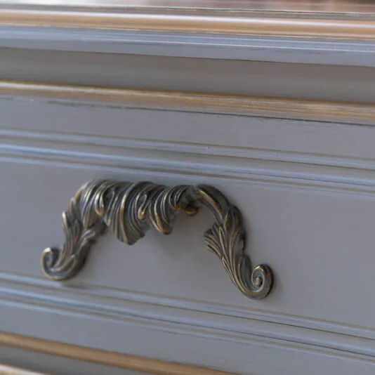 Tall Classic Louis Style Chest Of Drawers Close-up of a decorative handle on the Tall Classic Louis Style Chest of Drawers. The handle is ornately designed with leaf-like patterns, and the drawers are painted in a pale gray color with subtle gold accents.
