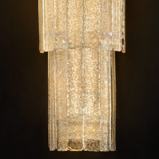 Large Tiered Amber Glass Wall Light A close-up of the Large Tiered Amber Glass Wall Light reveals its elegant, rectangular form with cascading layers of textured amber glass that seem to shimmer. This sophisticated lighting fixture emits a warm, ambient glow, accentuating the intricate details of the glass design against a dark background.