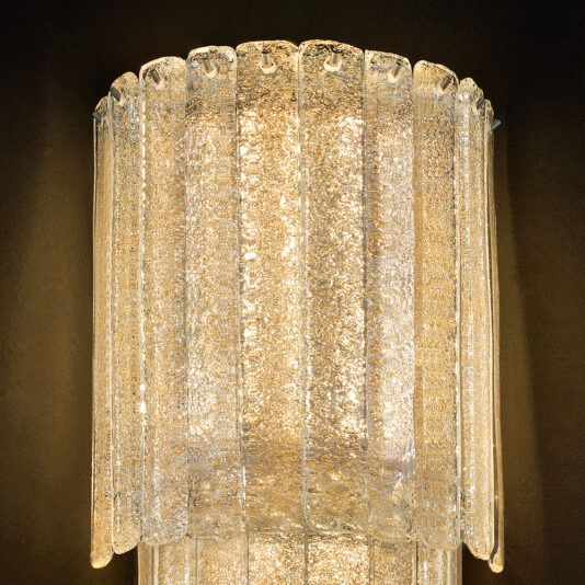Large Tiered Amber Glass Wall Light A close-up of the Large Tiered Amber Glass Wall Light reveals its elegant, modern cylindrical design with textured amber glass panels. The internal light source emits a warm, glowing light that accentuates the intricate details and texture of the large tiered glass design, all set against a dark background.