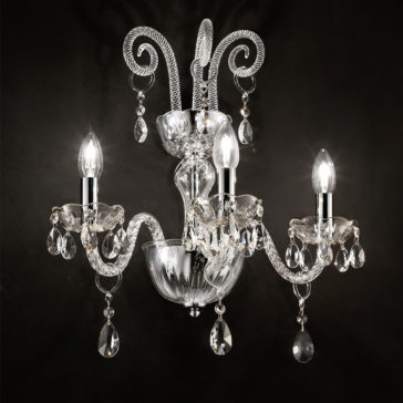 Triple Glass Wall Light With Crystal Drops The Triple Glass Wall Light With Crystal Drops features three ornate arms adorned with decorative crystal droplets, each holding a light bulb. Mounted against a dark background, its elaborate design includes intricately detailed curved arms and clear, shimmering glass elements.