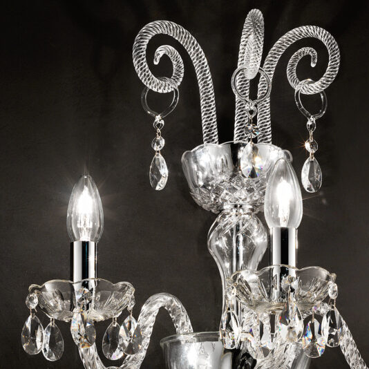 Triple Glass Wall Light With Crystal Drops A close-up of an elegant Triple Glass Wall Light With Crystal Drops showcases its intricate glasswork and crystal drops, with two lit candle-style bulbs illuminating the sophisticated and luxurious design against a dark background.