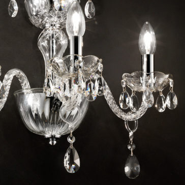 Triple Glass Wall Light With Crystal Drops Close-up of an elegant Triple Glass Wall Light With Crystal Drops with two lit bulbs. Each bulb is surrounded by ornate crystal decorations and dangling teardrop ornaments, mounted on a sleek chrome fixture against a dark background.