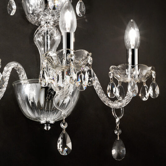 Triple Glass Wall Light With Crystal Drops Close-up of an elegant Triple Glass Wall Light With Crystal Drops with two lit bulbs. Each bulb is surrounded by ornate crystal decorations and dangling teardrop ornaments, mounted on a sleek chrome fixture against a dark background.
