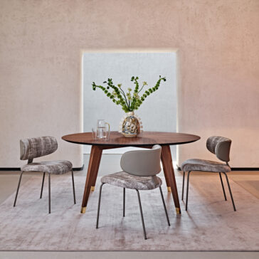 A modern dining room setup featuring the Contemporary Round Walnut Dining Table accompanied by three beige upholstered chairs. The table is adorned with a clear glass vase filled with green foliage and two drinking glasses. The background showcases a minimalist light-colored wall and a large window.
