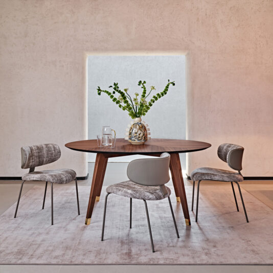 Contemporary Round Walnut Dining Table A modern dining room setup featuring the Contemporary Round Walnut Dining Table accompanied by three beige upholstered chairs. The table is adorned with a clear glass vase filled with green foliage and two drinking glasses. The background showcases a minimalist light-colored wall and a large window.