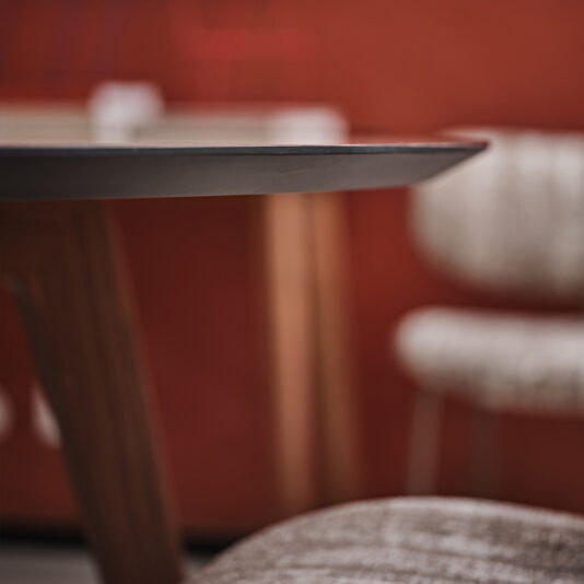 Contemporary Round Walnut Dining Table Close-up view of the Contemporary Round Walnut Dining Table, showcasing its sleek black surface and wooden legs. In the background, two cushioned chairs with textured upholstery are slightly blurred, set in front of a warm-toned wall.