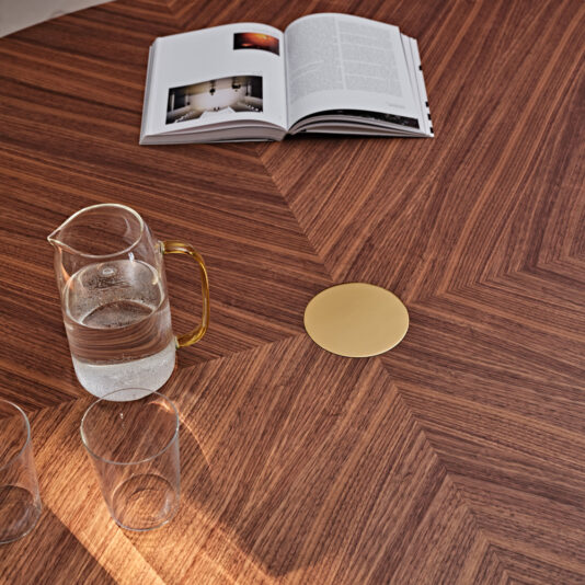 Contemporary Round Walnut Dining Table The Contemporary Round Walnut Dining Table with a geometric pattern showcases a glass pitcher of water and two empty glasses. Near the top of the image, an open book displaying photos and text can be seen. A small, golden object is placed at the center of the table's design.
