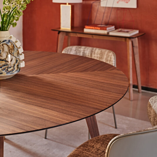 Contemporary Round Walnut Dining Table A modern dining room features the Contemporary Round Walnut Dining Table accompanied by upholstered chairs. A vase with white flowers decorates the table, while in the background, a console table displays books and geometric decor against a burnt orange accent wall.