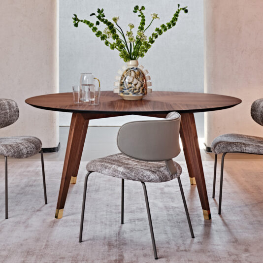 A contemporary dining room features the Contemporary Round Walnut Dining Table with sleek tapered legs and gold accents. Surrounding the table are four padded chairs with metal legs, and a decorative vase of green and white flowers serves as an elegant centerpiece. The room maintains a light, neutral palette.