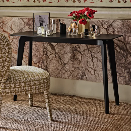 A stylish vanity setup with a Contemporary Ash Mocha Console Table against a marbled wall showcases framed photos, perfume bottles, red flowers in a vase, and cosmetics. A patterned fabric cushioned chair sits in front on a textured carpet.