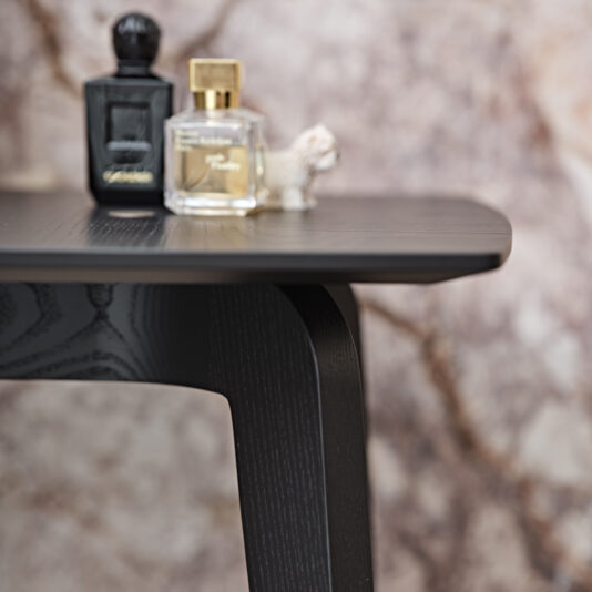 A close-up of a Contemporary Ash Mocha Console Table, displaying its modern black wooden design, with two perfume bottles and a small decorative figurine adorning the top. The background is blurred with earthy tones, providing a soft contrast to the sharp lines of this stylish piece.