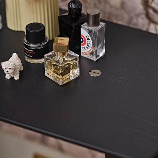A Contemporary Ash Mocha Console Table holds a variety of items, including a small white dog figurine, a bottle of Le Labo Santal 33 perfume, a square glass perfume bottle with a gold cap, an Engelskt Gin bottle, and a yellow ribbed container.
