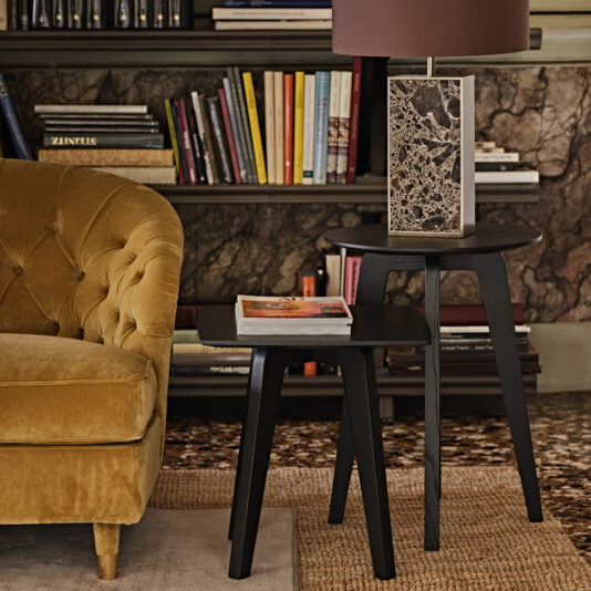 A cozy living room corner featuring a mustard velvet tufted sofa, a Contemporary Mocha Ash Side Table with a book and magazine on top, a lamp with a marble base, and a bookshelf filled with colorful books against a marble-patterned wall.