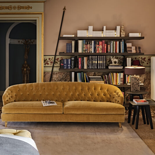 A cozy, elegant living room features a mustard yellow tufted sofa, a Contemporary Mocha Ash Side Table with a lamp, and a colorful, well-organized bookshelf against a decorated wall. The room has warm, inviting tones and intricate design details on the walls and arches.