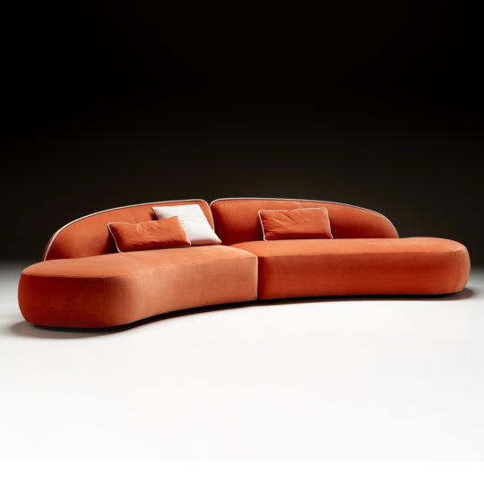 Set against a dark background, the Contemporary Chaise Style Modular Sofa features a modern, curved orange design with a smooth, rounded aesthetic. This sleek and minimalist piece boasts seamless plush upholstery and comes with four matching orange cushions as well as one contrasting white cushion.