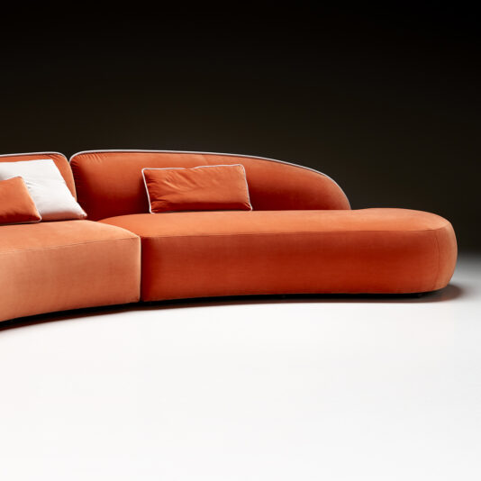 Contemporary Chaise Style Modular Sofa Image of the Contemporary Chaise Style Modular Sofa, featuring a modern, curved design in vibrant orange with soft, rounded edges. The sofa is placed against a dark background and includes two matching orange cushions paired with one contrasting white cushion. A sleek white floor beneath enhances the minimalistic and stylish contrast.