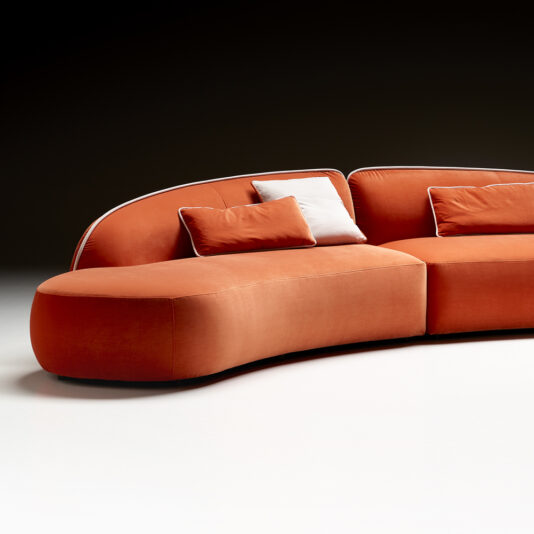 Contemporary Chaise Style Modular Sofa A modern Contemporary Chaise Style Modular Sofa with a sleek, curved design, upholstered in vibrant orange fabric. This contemporary seating features plush cushioning and is adorned with two orange rectangular pillows and a white square pillow. The backdrop is a simple dark gradient, highlighting the sofa's vibrant color.